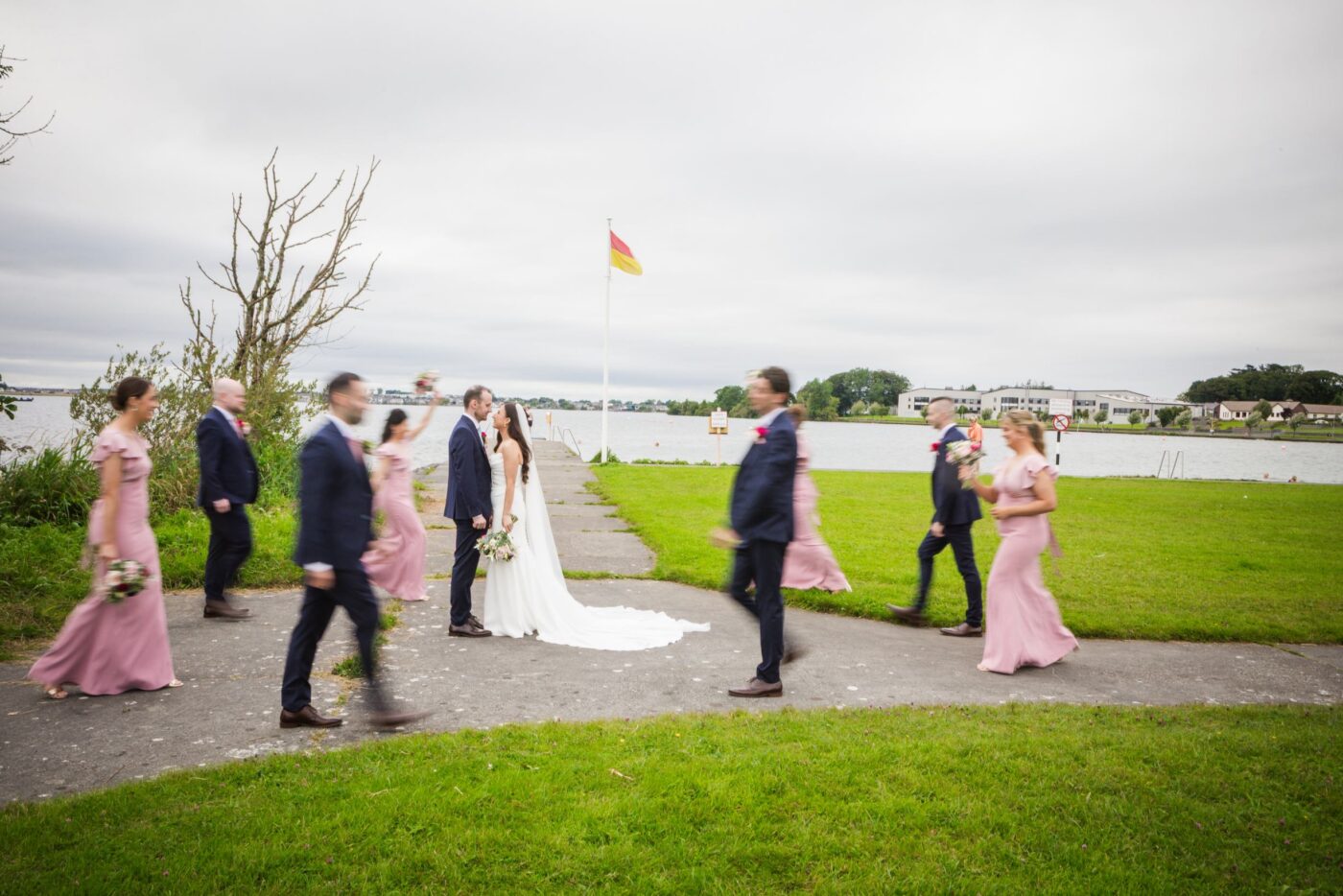 Bride and groom facing each other beside a lake as bridesmaids in pink and groomsmen in navy walk around them.