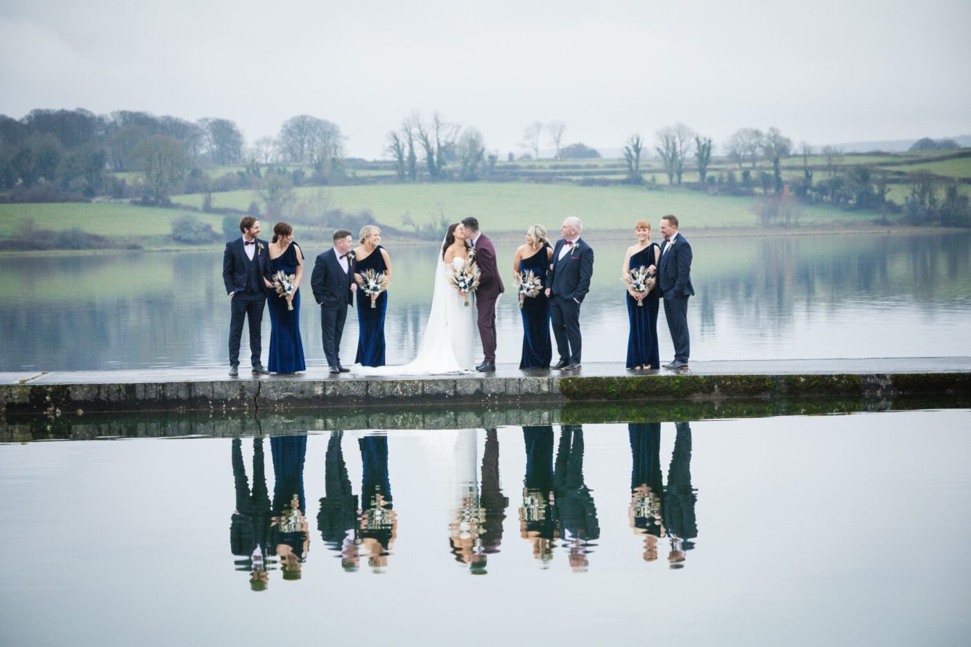 Bride and groom kissing between their wedding party on a pier, with the group reflected in the calm waters of Loughrea Lake.