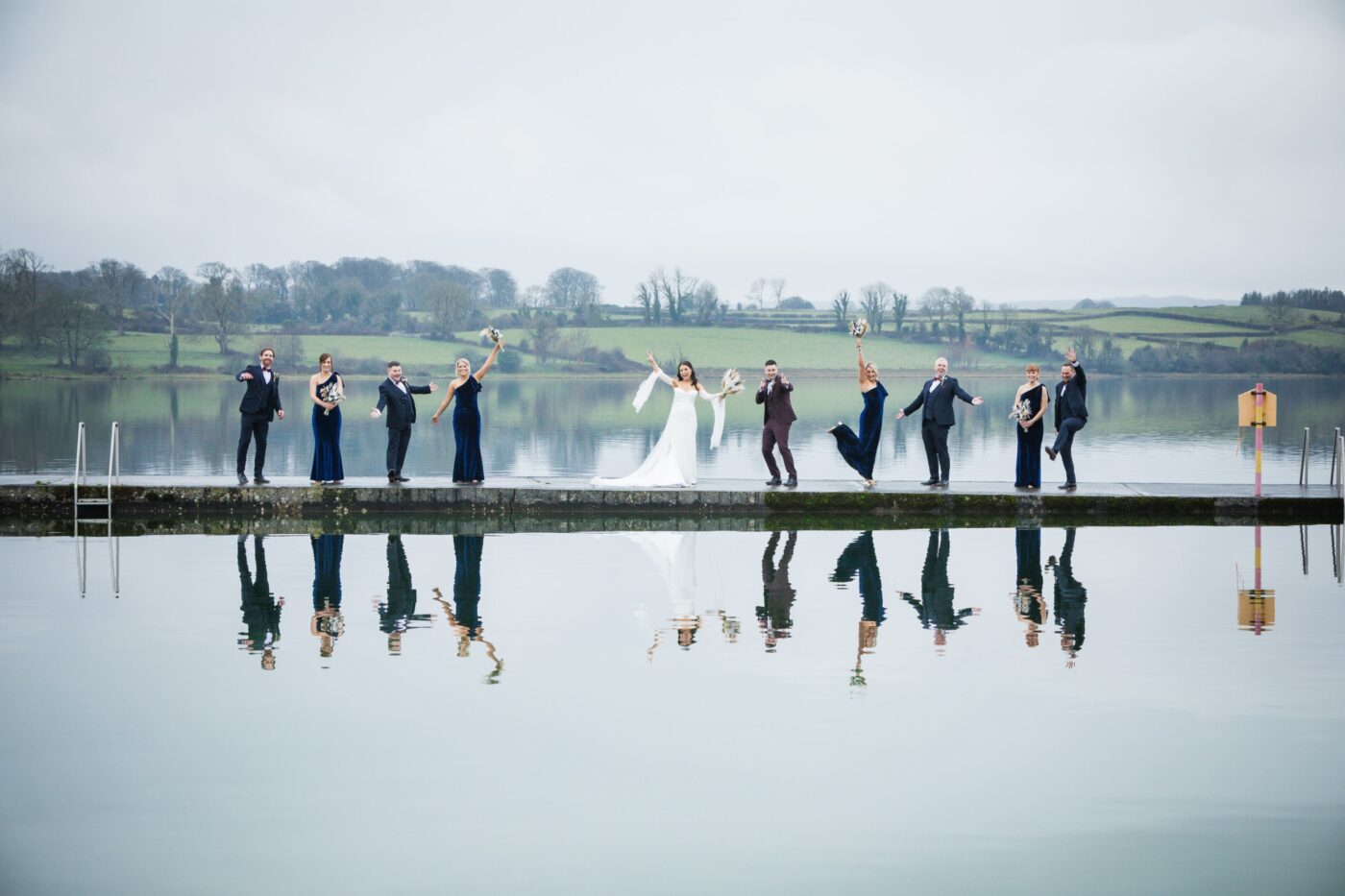 Wedding party raising their arms and bouquets along a pier at Loughrea Lake, with their reflections in the water.