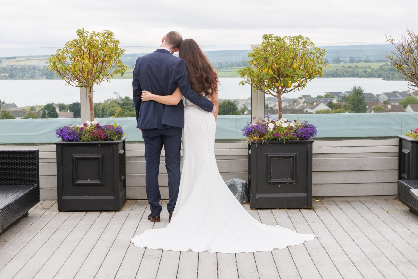 Bride and groom seen from behind, embracing between potted trees on the rooftop terrace at Loughrea Hotel & Spa overlooking Loughrea Lake.