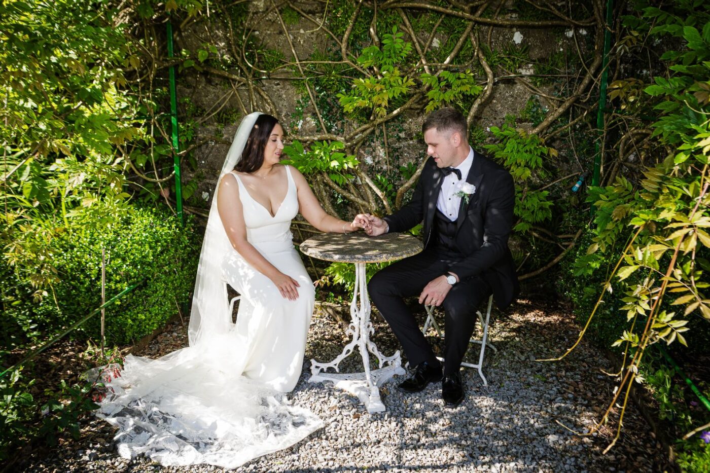 Bride and groom seated at a small garden table, holding hands beneath climbing greenery.