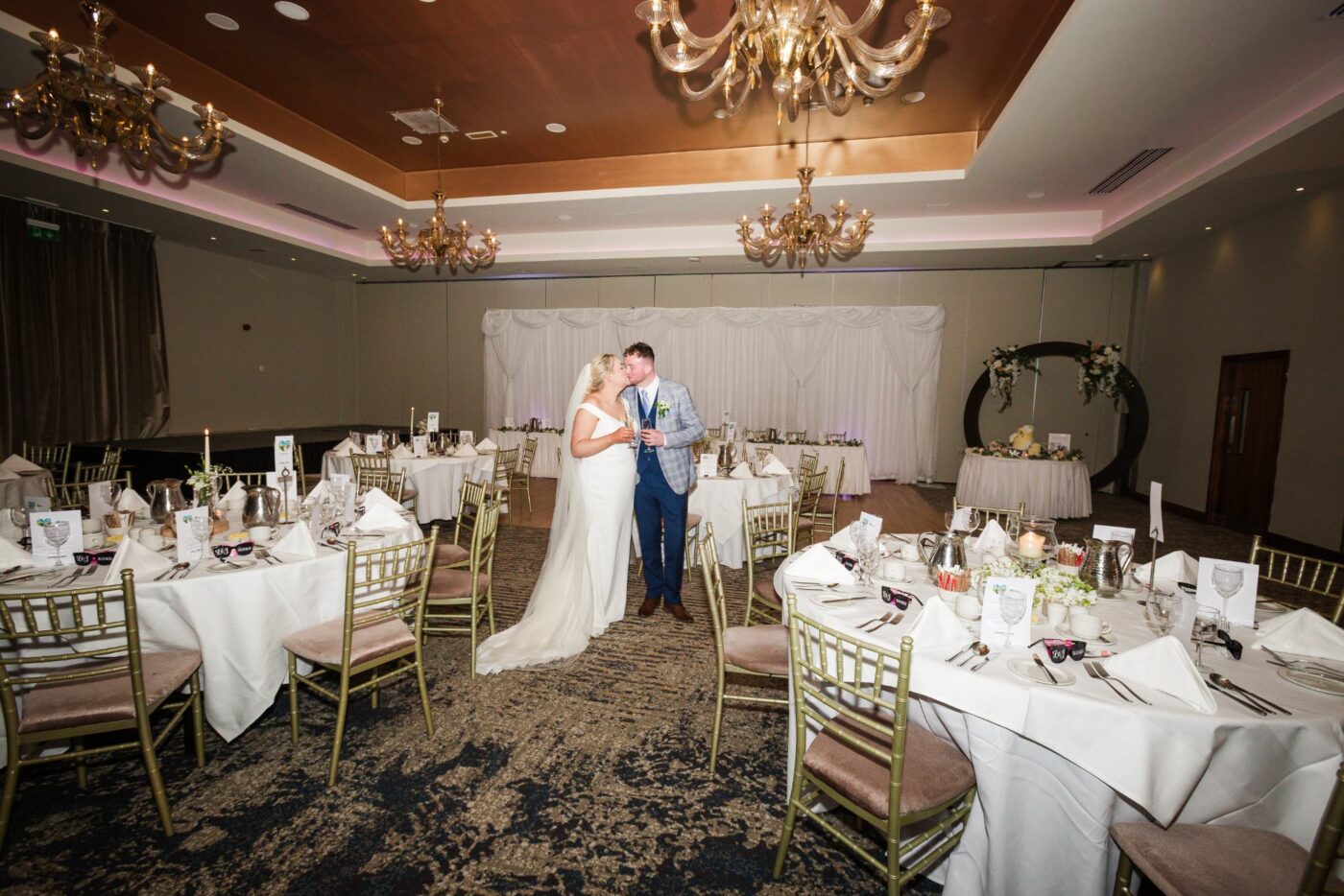 Bride and groom kissing with drinks in hand among dressed reception tables beneath chandeliers at Loughrea Hotel & Spa.