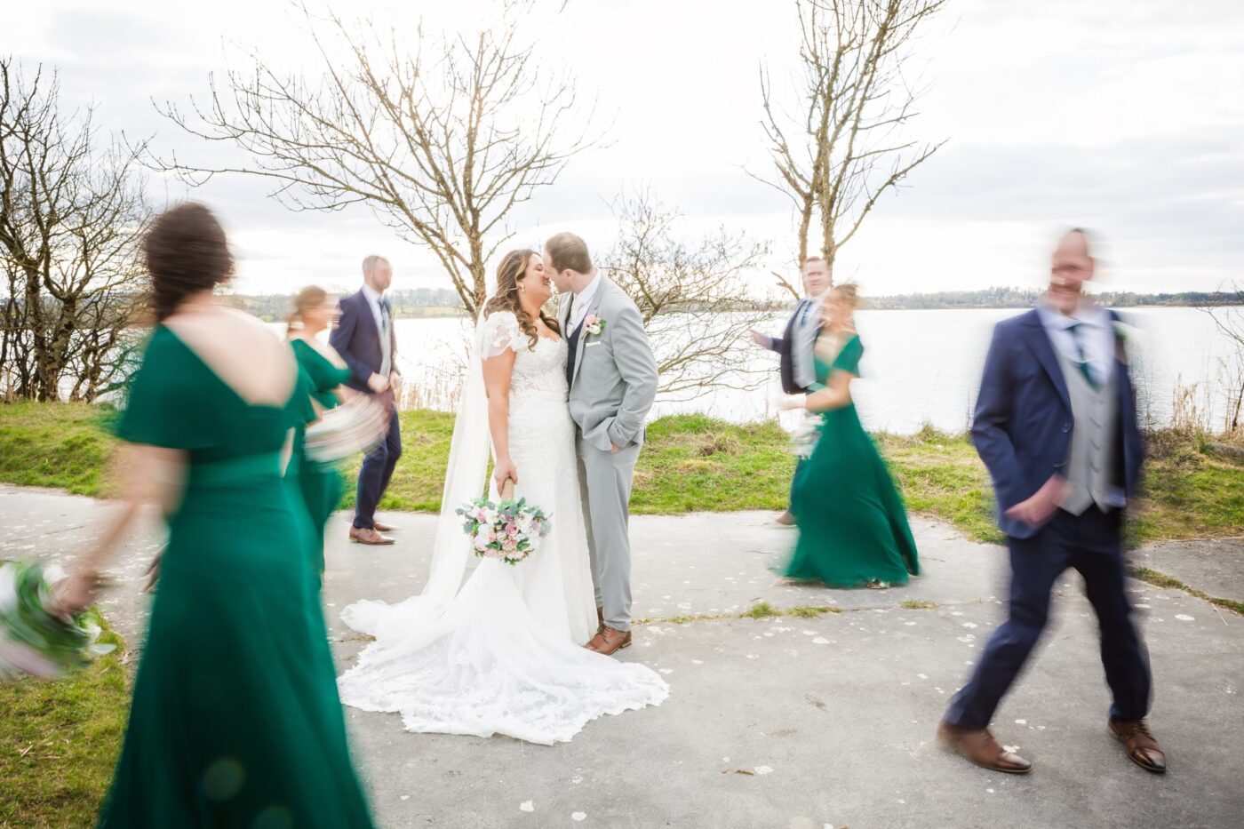 Bride and groom kissing beside Loughrea Lake as bridesmaids in emerald green and groomsmen walk around them in motion blur.