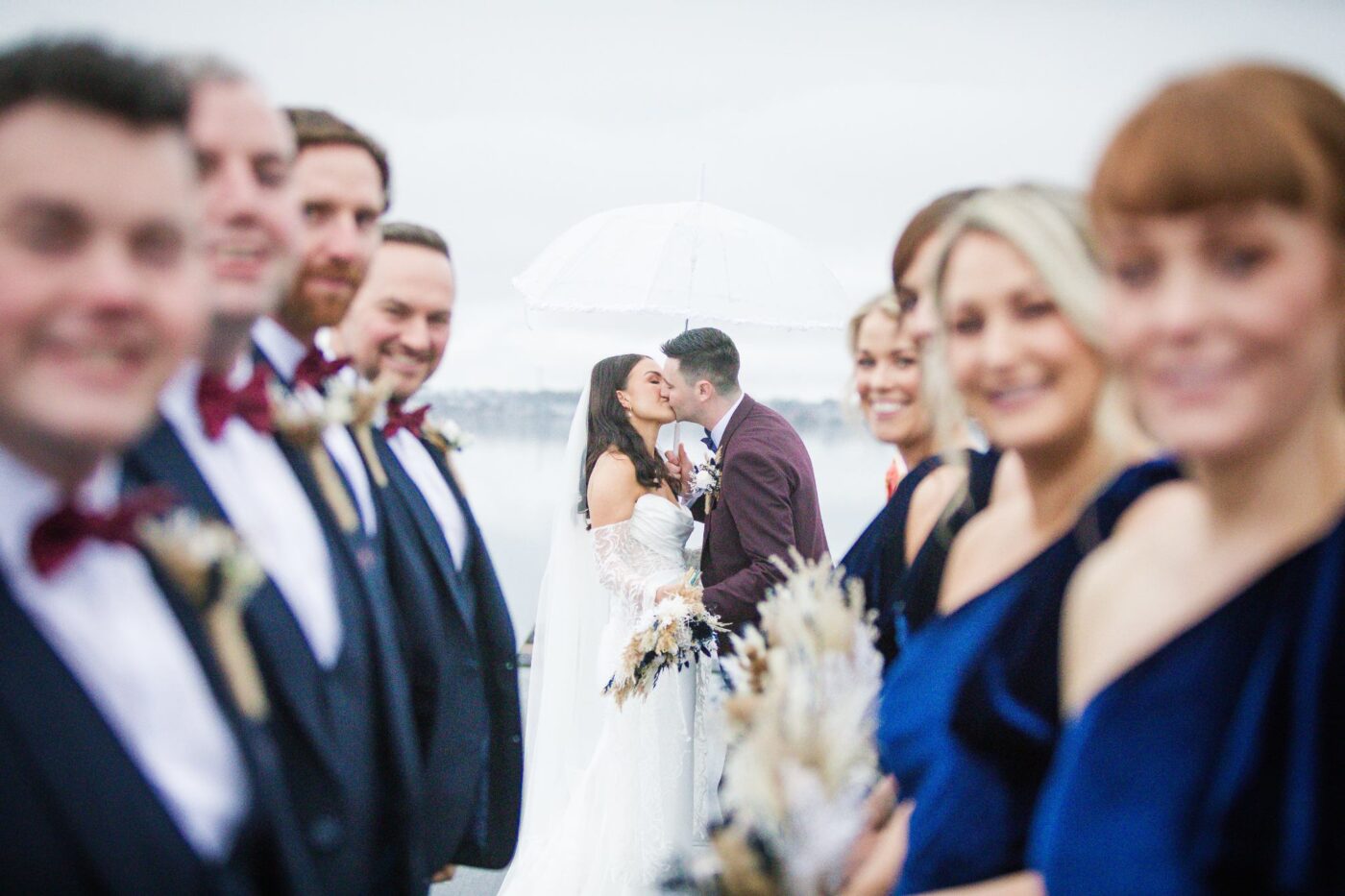 Bride and groom kissing beneath a clear umbrella, framed by softly blurred bridesmaids and groomsmen.