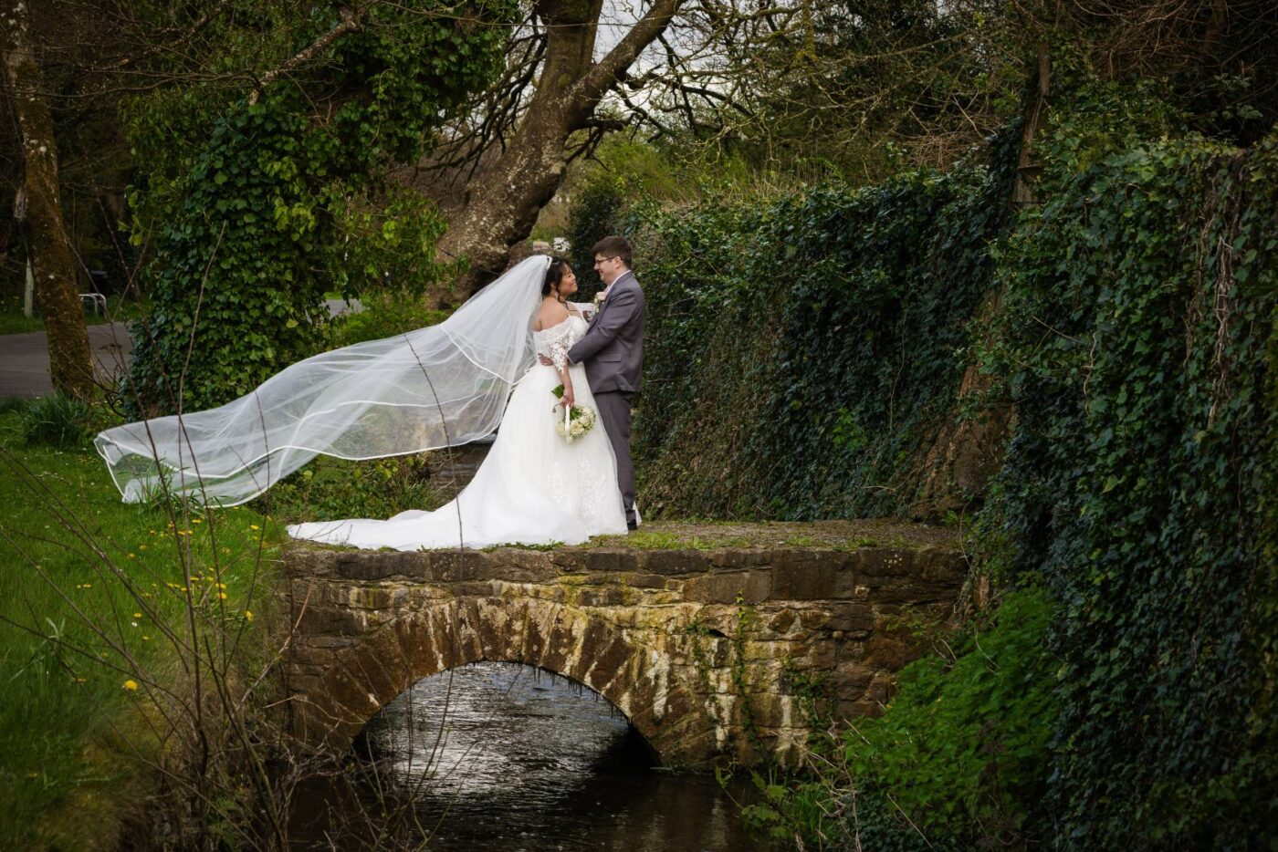 Bride and groom embracing on a stone bridge over a stream, with her long veil blowing to the side.