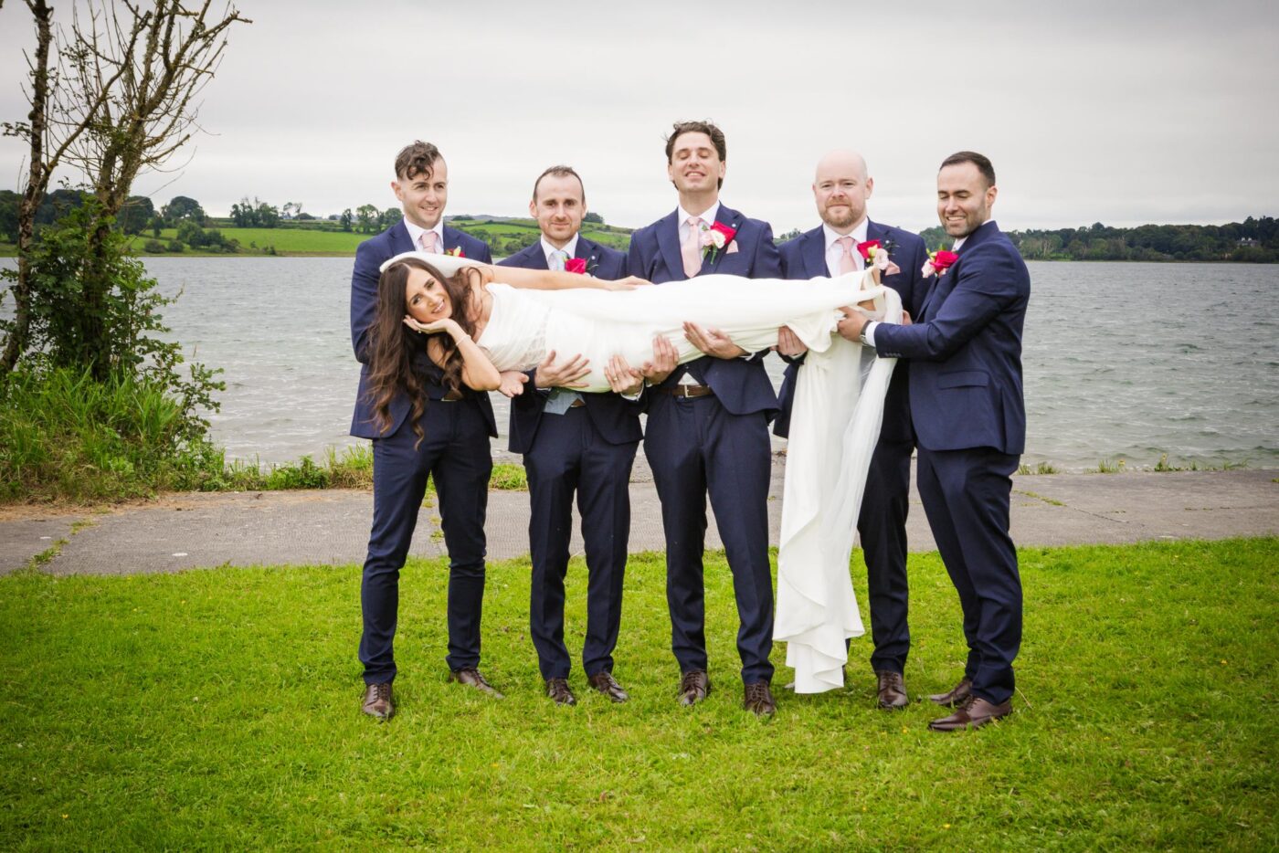 Smiling bride held horizontally by five men in navy suits and pink ties on grass beside Loughrea Lake.