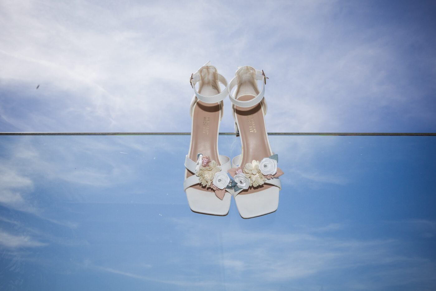 White bridal sandals with pastel floral decorations resting on a glass railing against a blue sky.