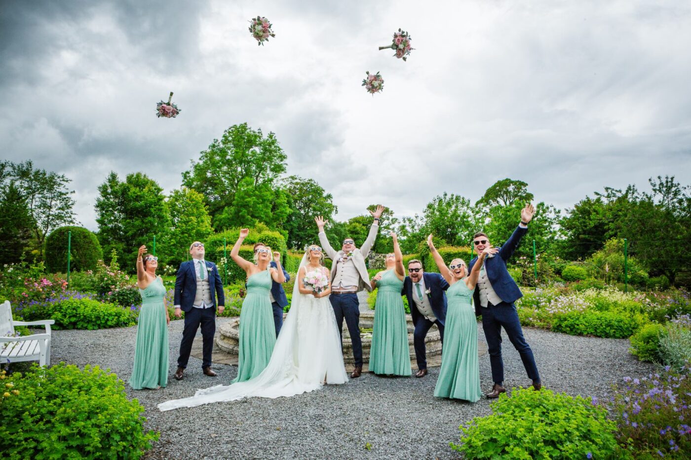 : Bridesmaids tossing their bouquets into the air as the bride, groom and wedding party cheer in a garden.