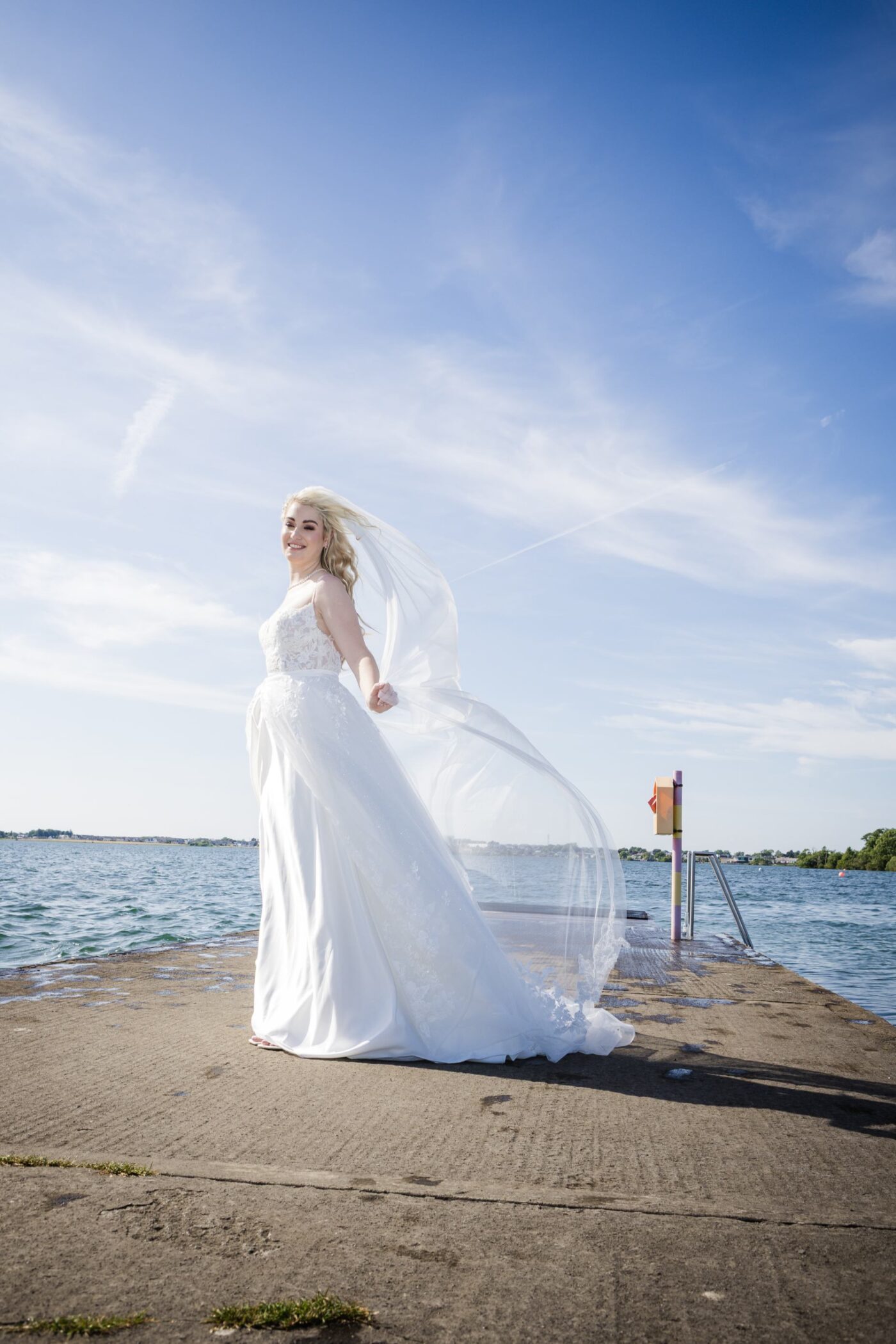 Smiling bride on a pier at Loughrea Lake beneath a blue sky, with her long veil blowing behind her.