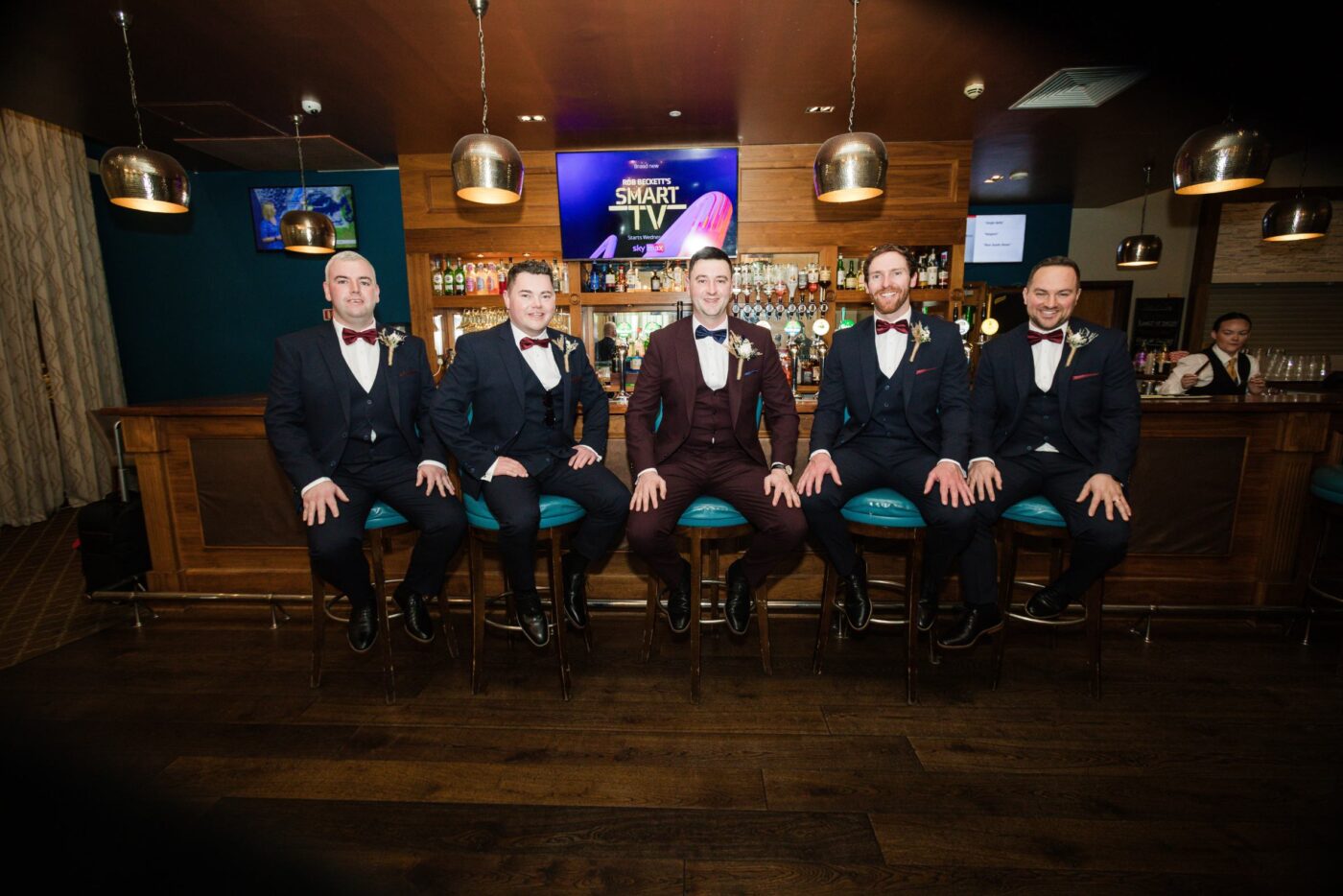 Groom in a burgundy suit seated on a bar stool at Loughrea Hotel & Spa with four groomsmen in navy suits and burgundy bow ties.