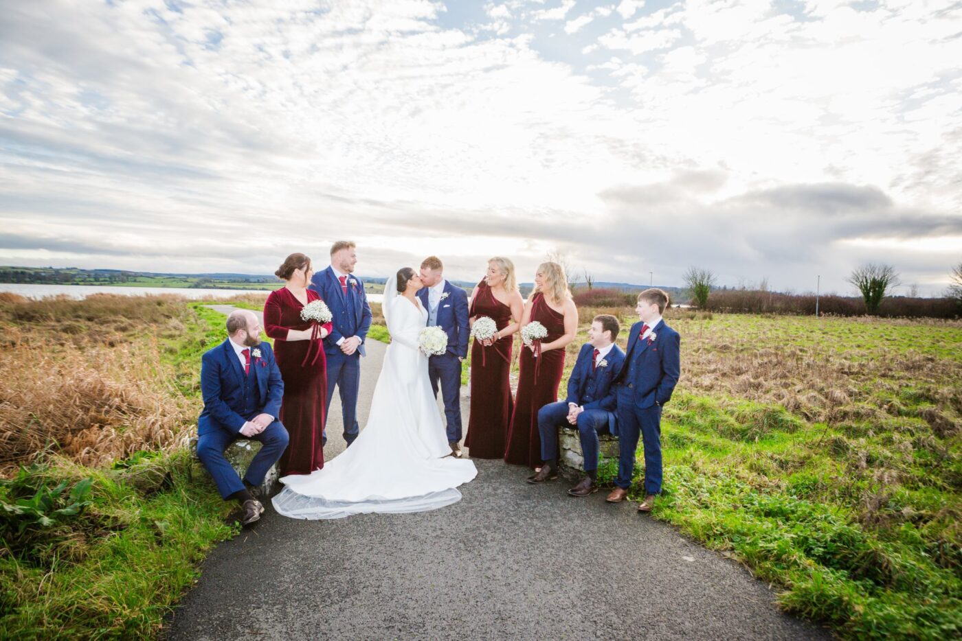 Bride and groom kissing on a lakeside path, surrounded by bridesmaids in burgundy and groomsmen in navy.