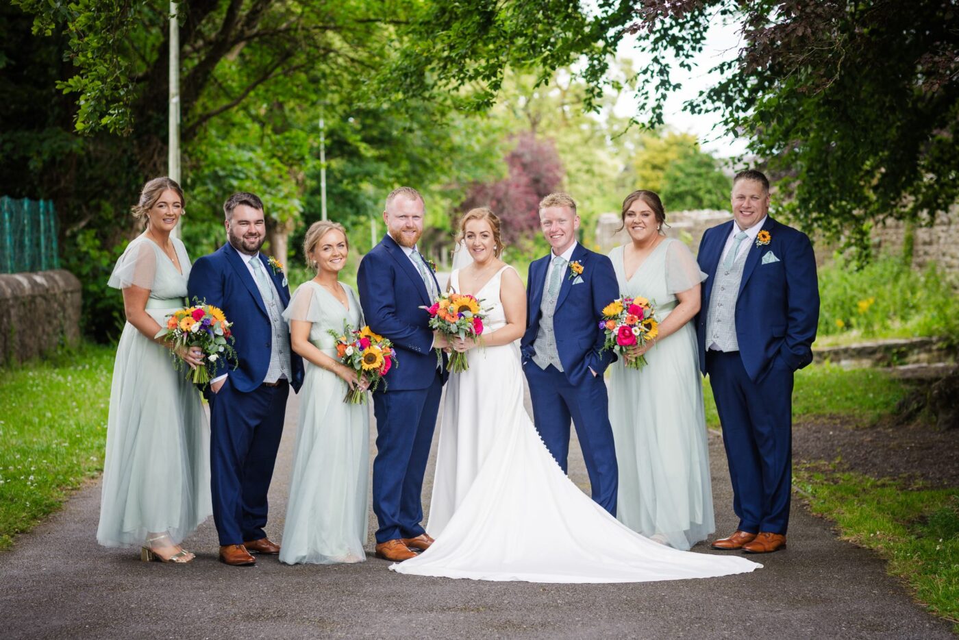 Bride and groom with bridesmaids in pale green and groomsmen in navy on a tree-lined path in Loughrea, holding colourful sunflower bouquets.