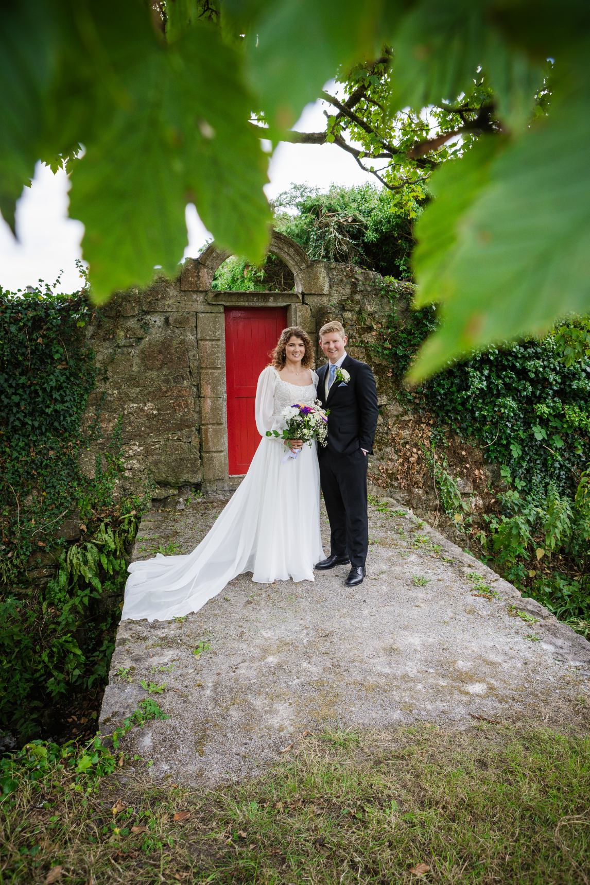 Bride and groom posing beside a red door in an old stone wall, framed by green leaves.