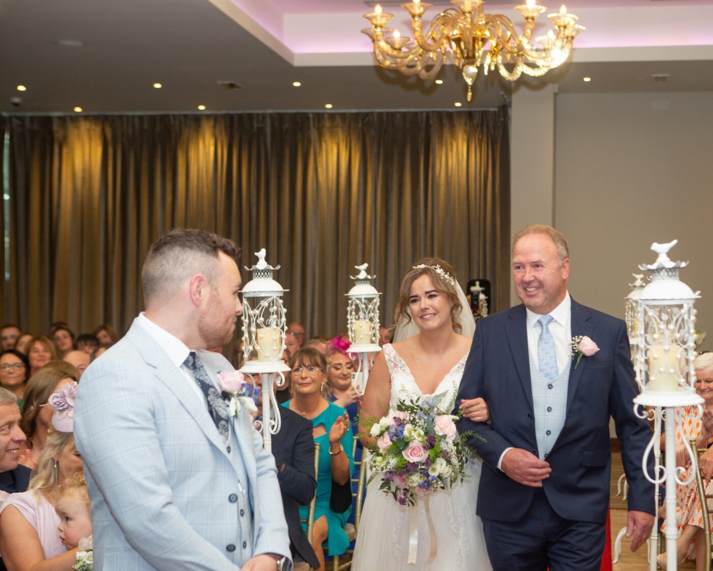Bride walking arm in arm with a man in a navy suit towards the groom, between white lanterns and seated guests.