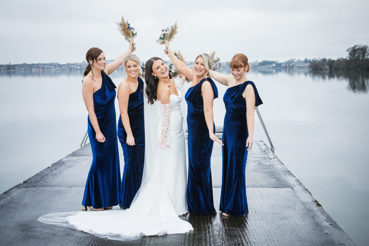 Laughing bride with four bridesmaids in navy velvet dresses raising bouquets on a pier at Loughrea Lake.