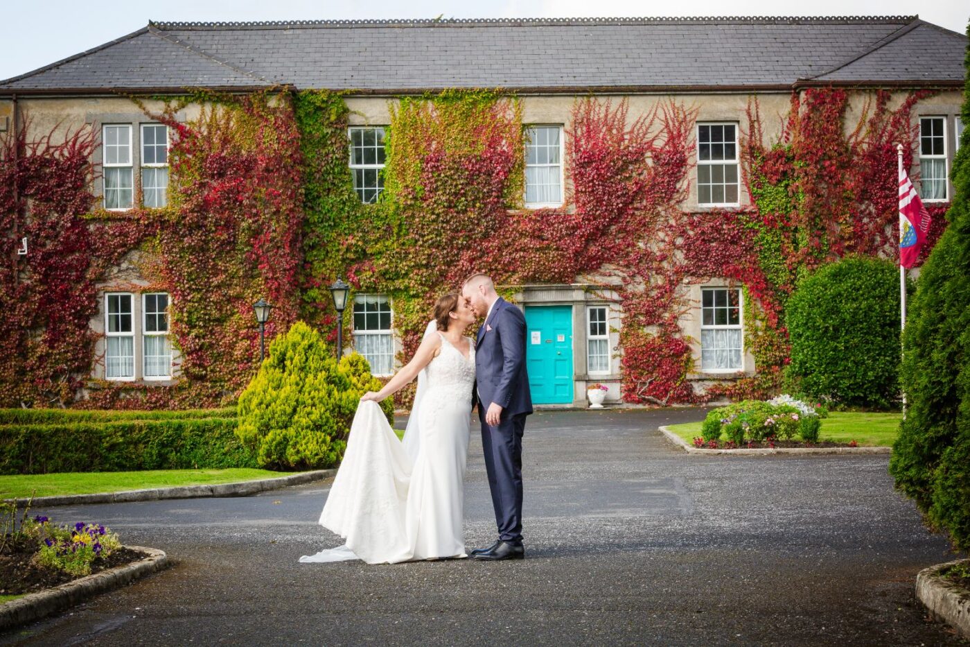 Bride and groom kissing outside a building covered in red and green ivy, with turquoise doors behind them.