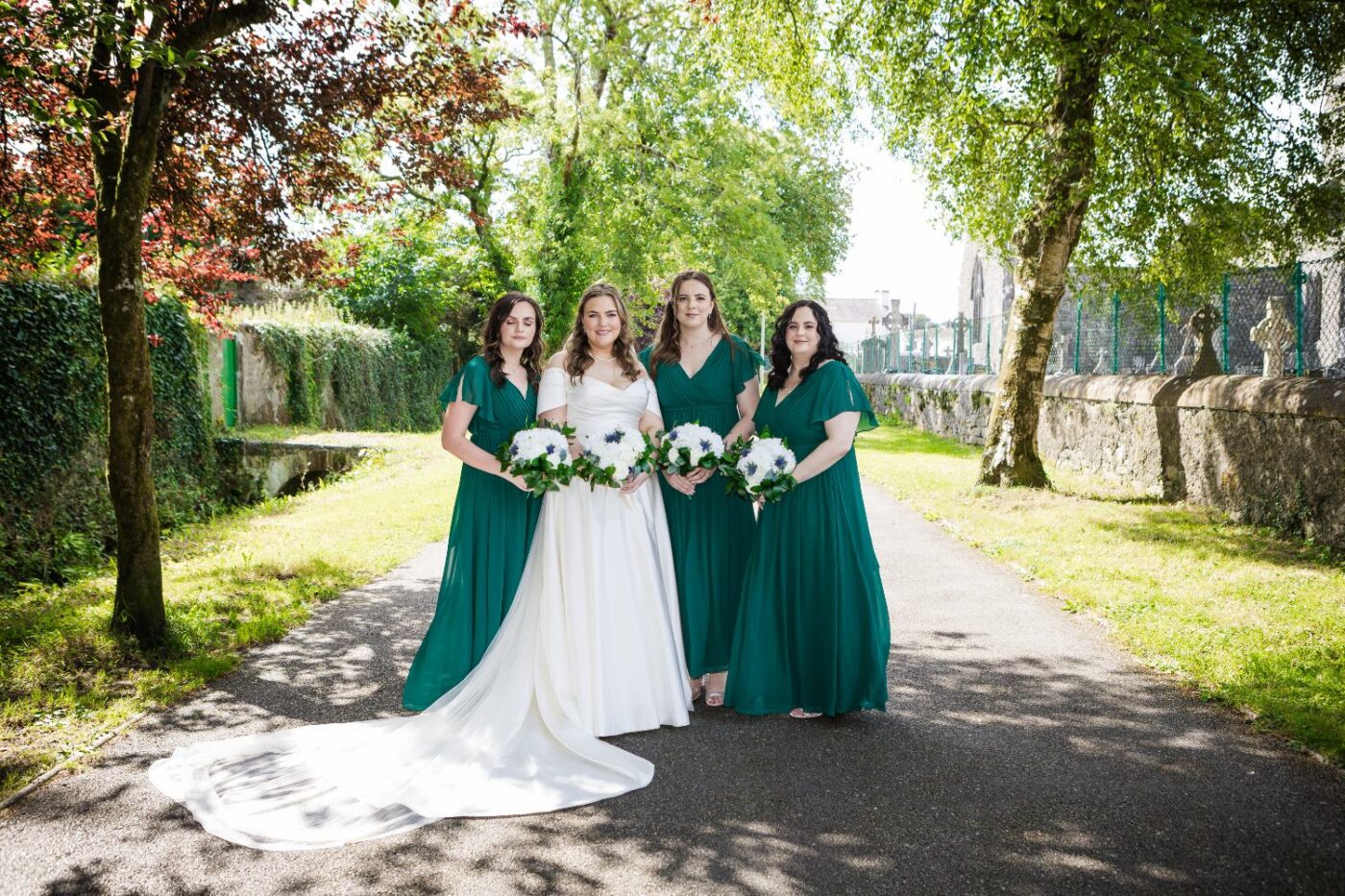 Bride with three bridesmaids in emerald green holding white bouquets on a sunny tree-lined path in Loughrea.