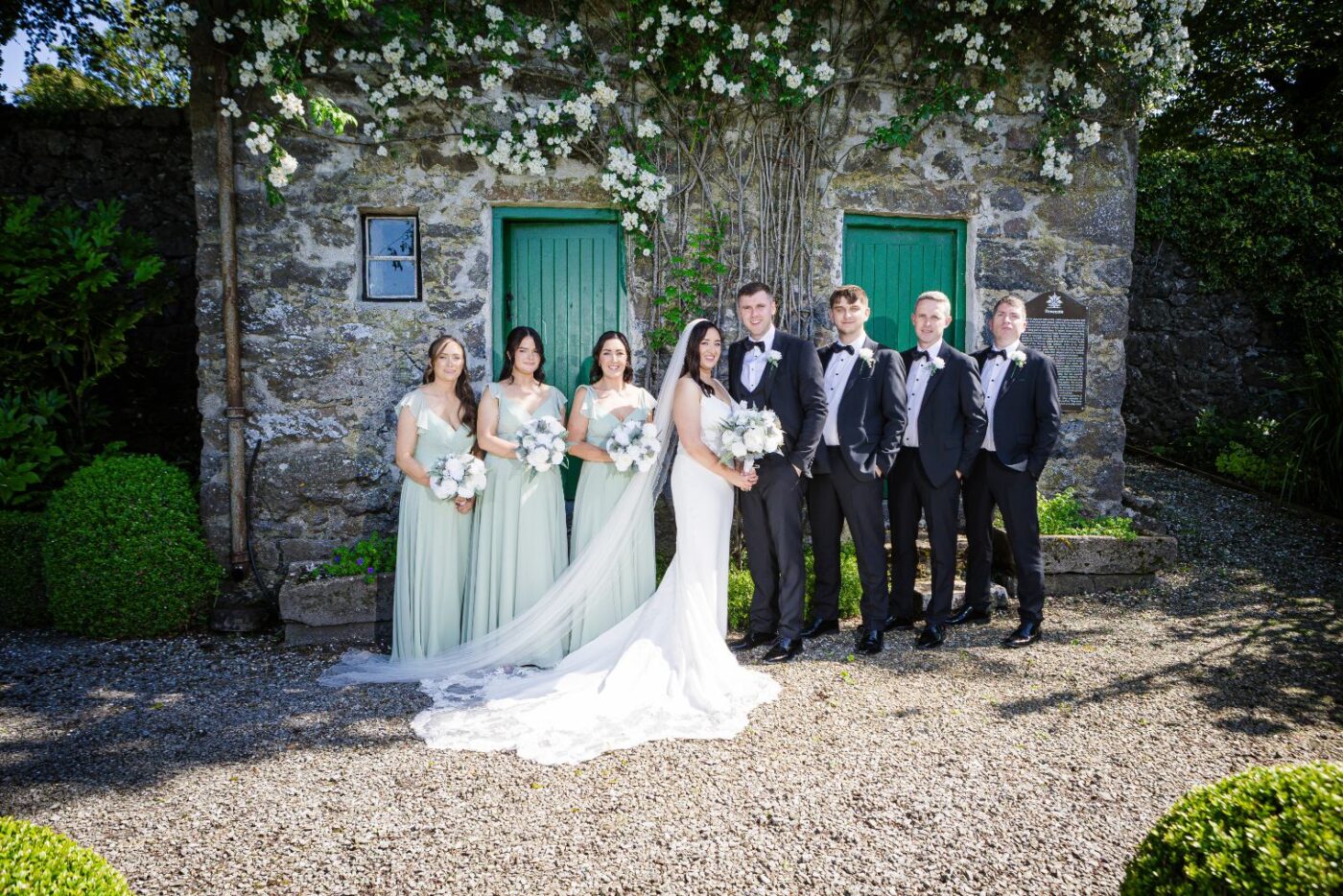 Bride and groom with bridesmaids in pale green and groomsmen in black outside a stone building with green doors and white climbing flowers.