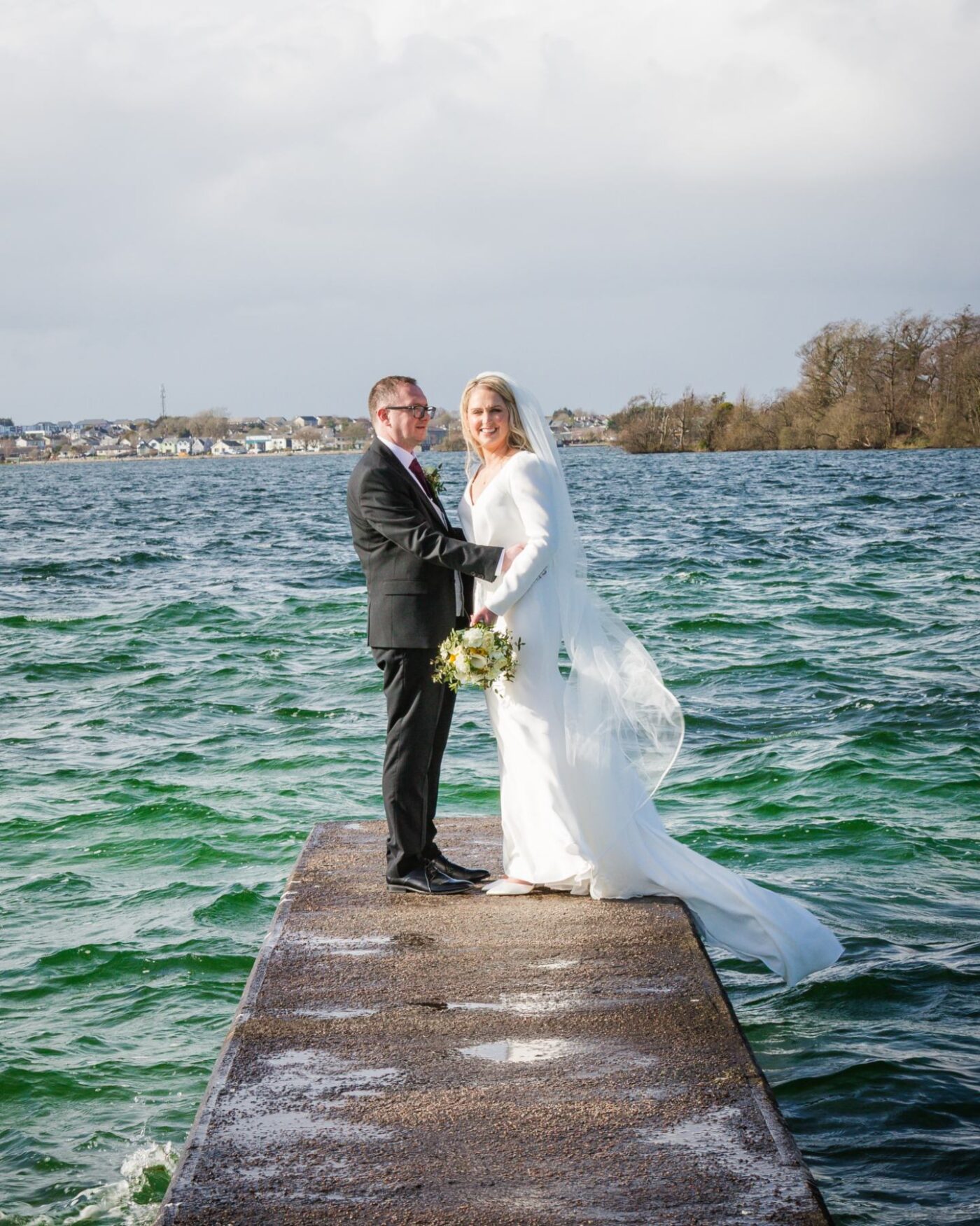 Bride and groom embracing on a pier beside the choppy waters of Loughrea Lake, with her veil blowing to the side.