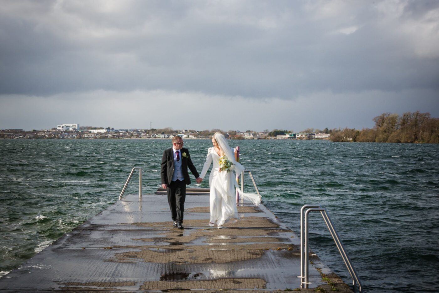 Bride and groom walking hand in hand along a wet pier at Loughrea Lake beneath dark clouds.
