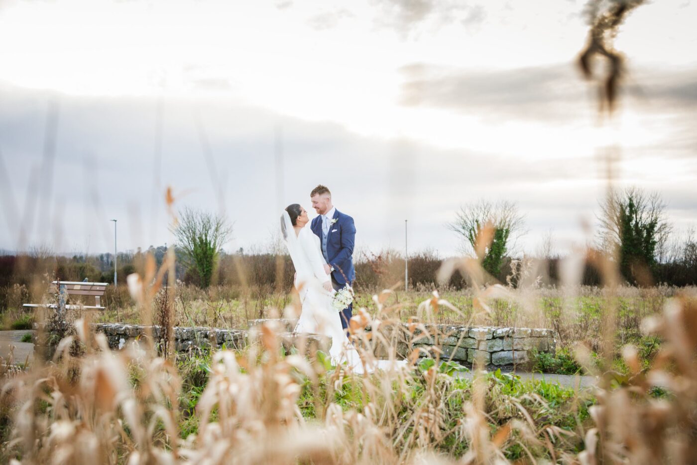 Bride and groom smiling at each other beside a low stone wall, framed by softly blurred wild grasses.