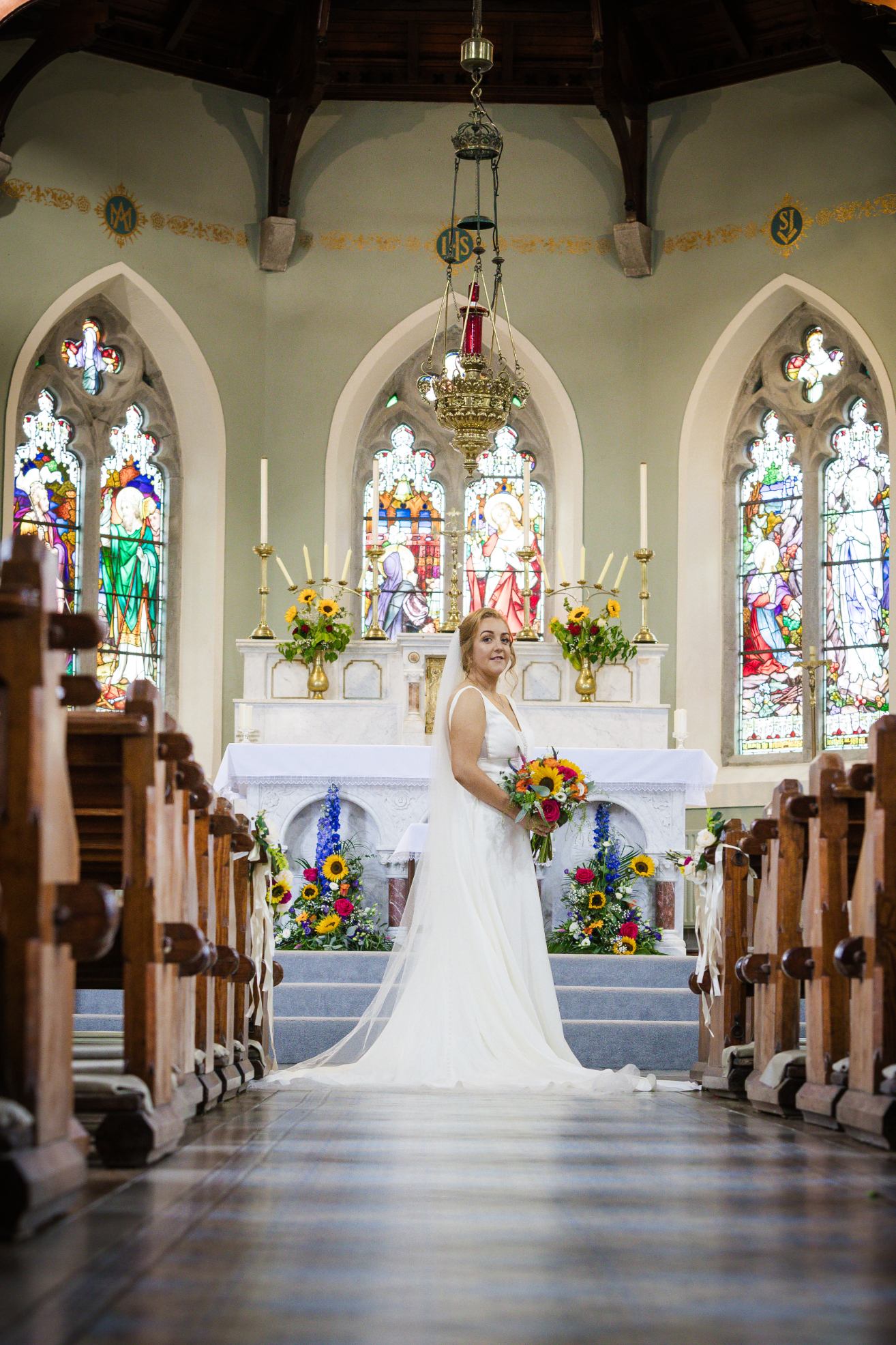 Bride holding a colourful sunflower bouquet in front of a church altar and stained-glass windows.