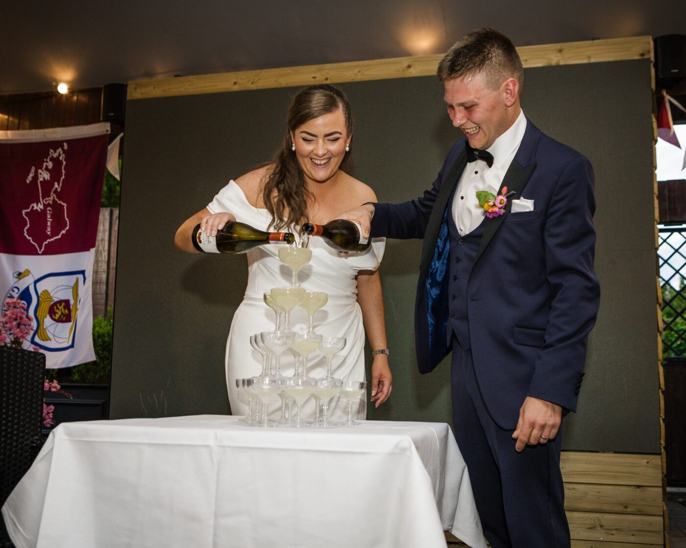 Laughing bride and groom pouring sparkling wine together into the top glass of a champagne tower.