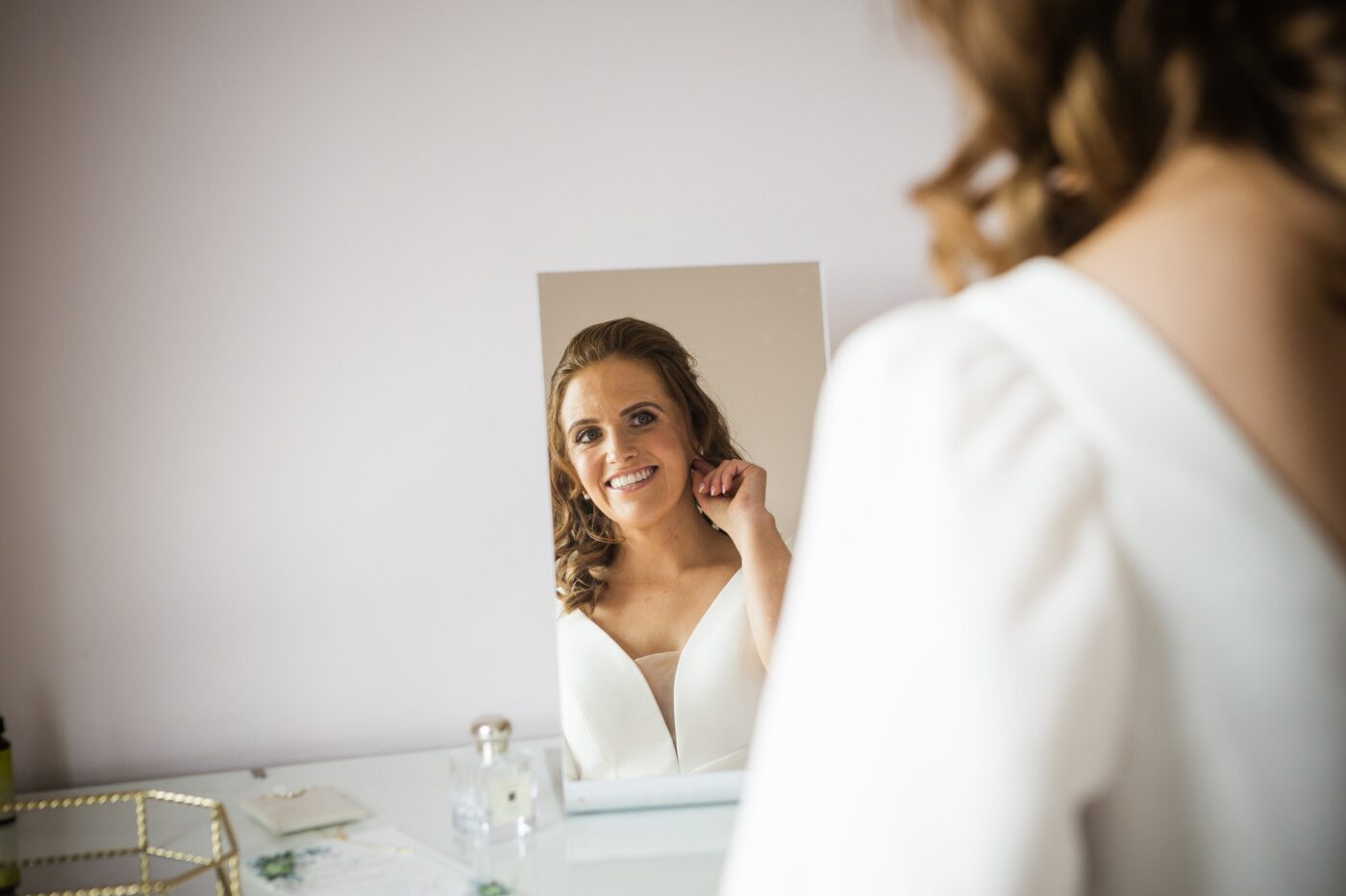 Smiling bride adjusting her earring while looking into a mirror.