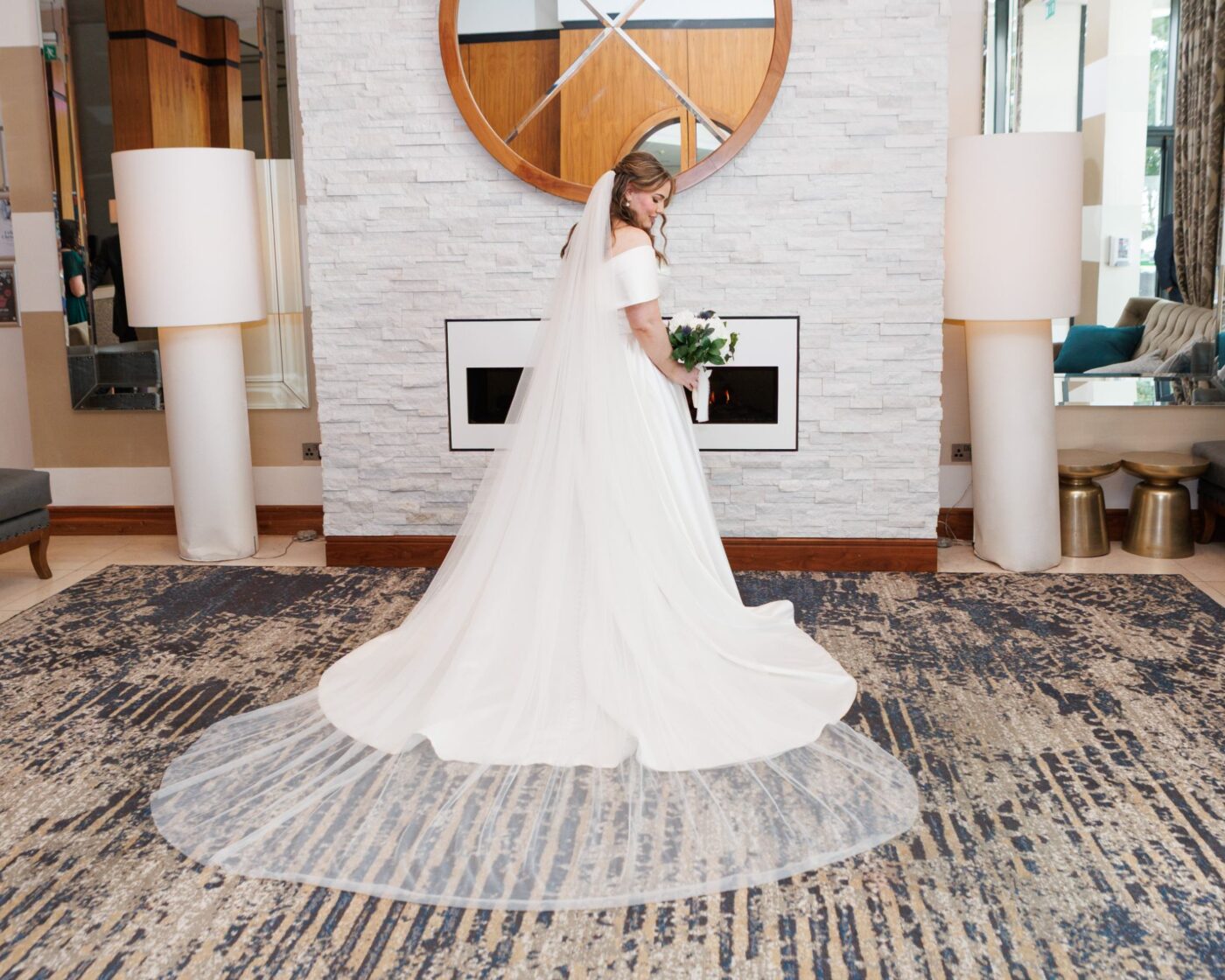 Bride holding a white bouquet beside a hotel fireplace, with her dress train and veil spread across the carpet.