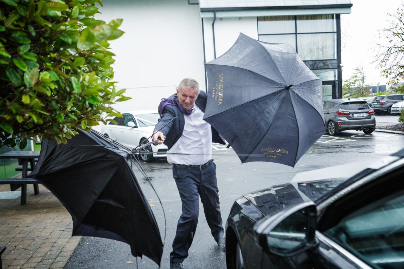 Man struggling with two windblown umbrellas outside a hotel beside a parked car.
