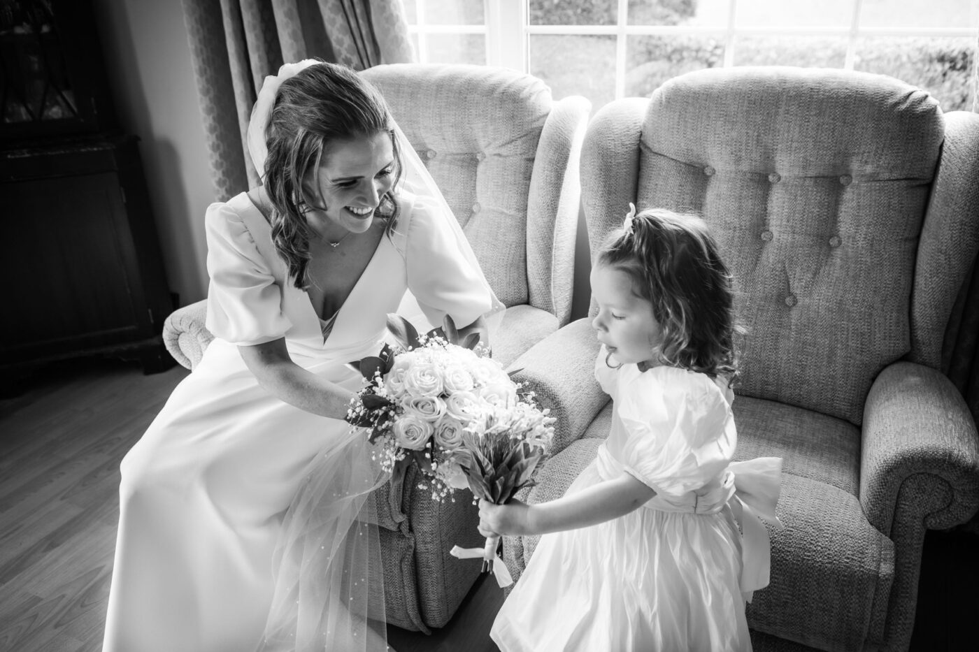 Black and white photograph of a bride smiling at a flower girl as they hold bouquets beside a window.