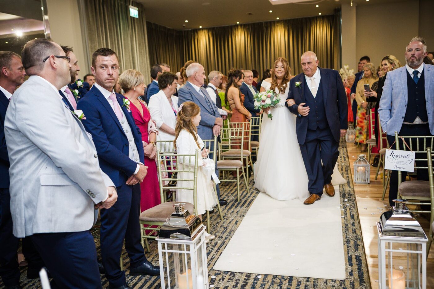 Bride walking arm in arm with a man in a navy suit along a lantern-lined aisle as wedding guests stand.